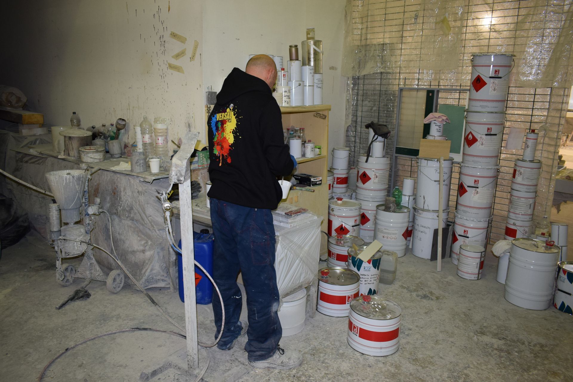 Un uomo con una felpa nera con cappuccio è in piedi in un laboratorio disordinato, pieno di barattoli di vernice e attrezzature; l'ambientazione è al chiuso.