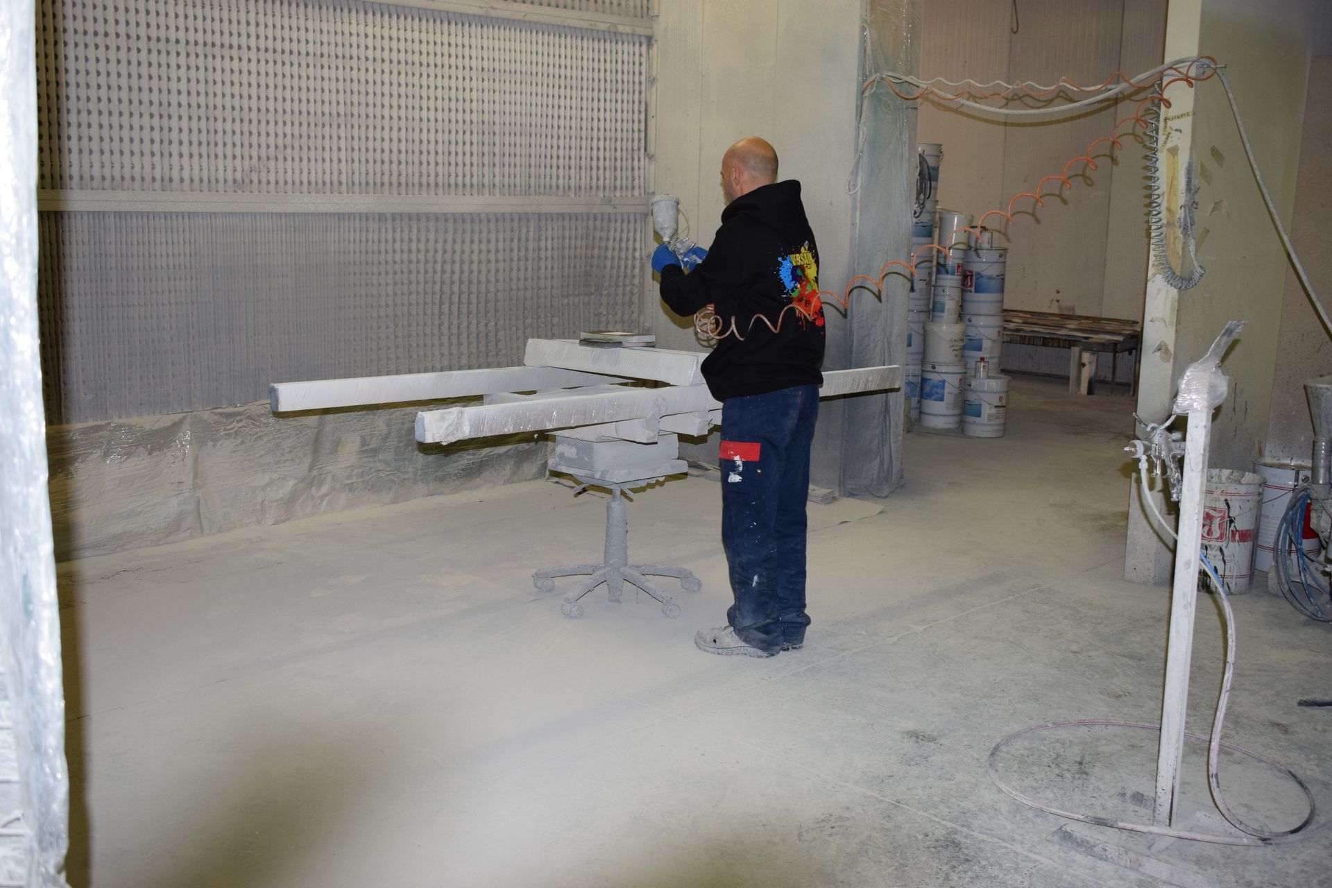 Persona che spruzza vernice sugli oggetti in una cabina di verniciatura. La vernice bianca ricopre gli oggetti e il pavimento.