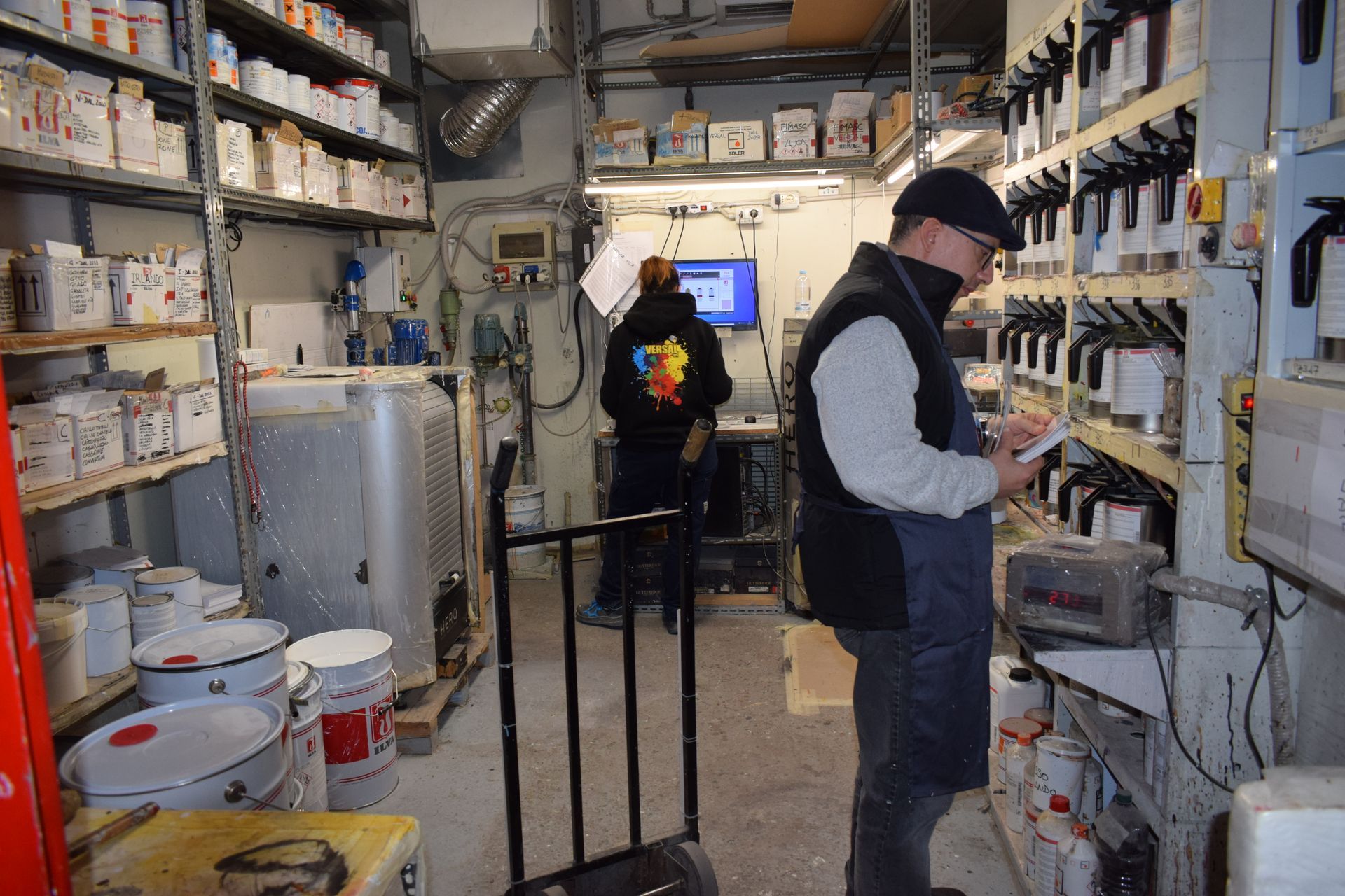 Due persone nella stanza dove si mescolano i colori, una sta scrivendo, scaffali pieni di barattoli di vernice.