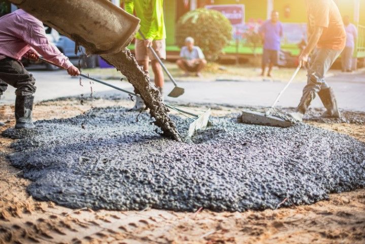 Workers pour wet concrete from a truck chute onto a construction site while others use tools to level the mixture.