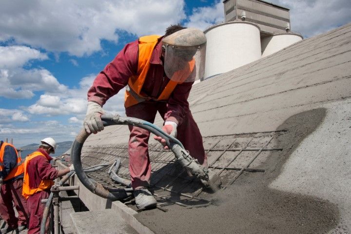 Two construction workers in orange safety vests and face shields apply wet concrete to a sloped surface with a hose.