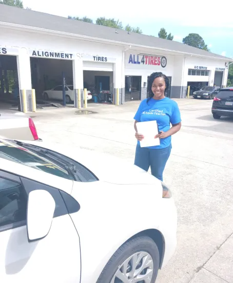 Person in blue shirt holding paper beside a white car outside an All Tire & Alignment shop. | All 4 Tires