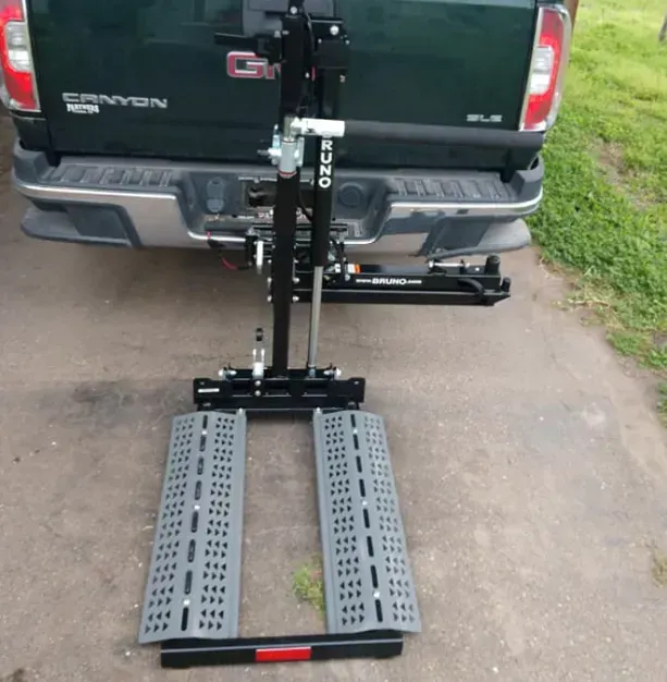 Wheelchair lift attached to the back of a green GMC truck. Ramp is down on a driveway.