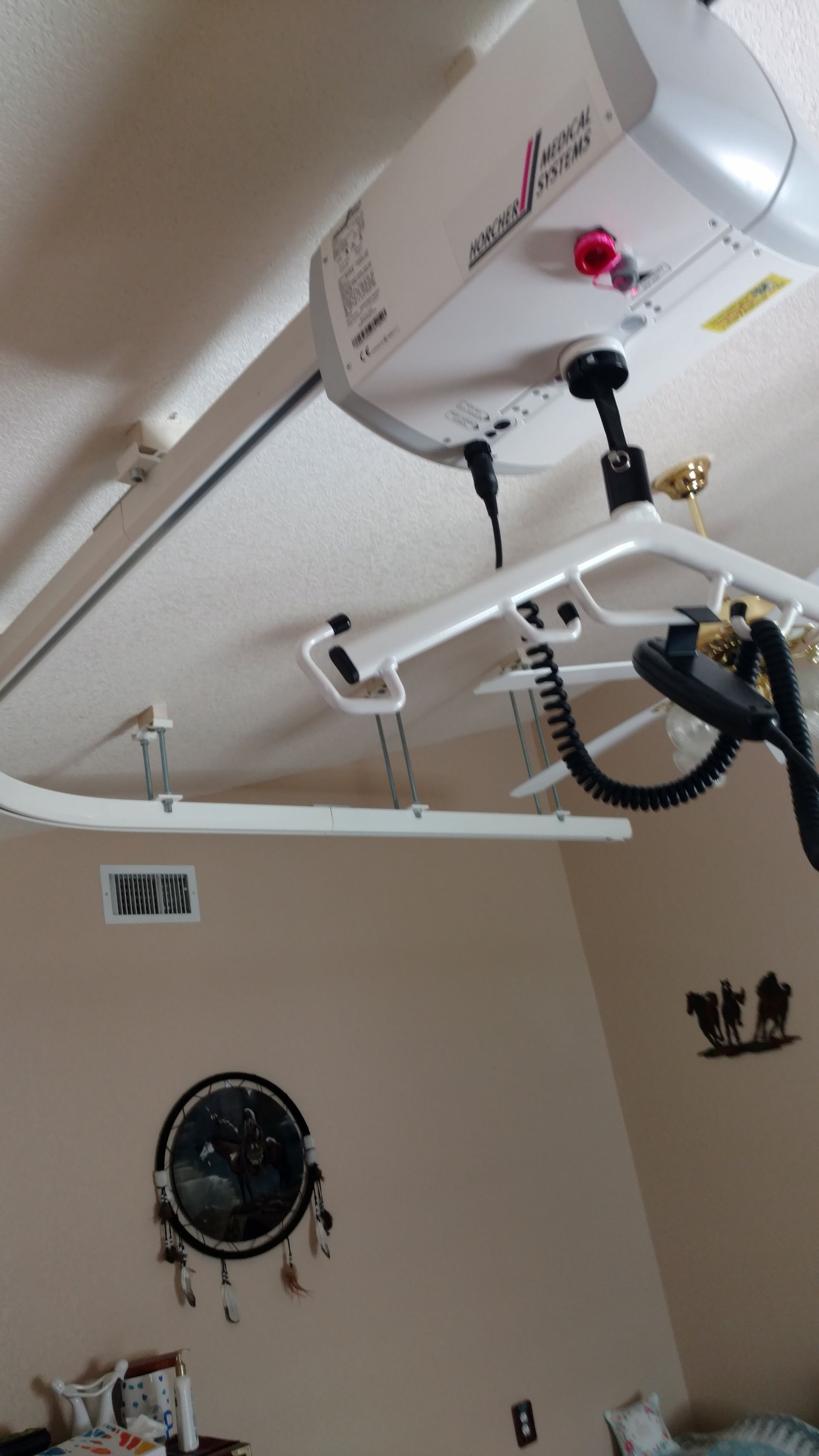 Ceiling-mounted patient lift system in a bedroom. White track and lift unit with a black control arm and harness.