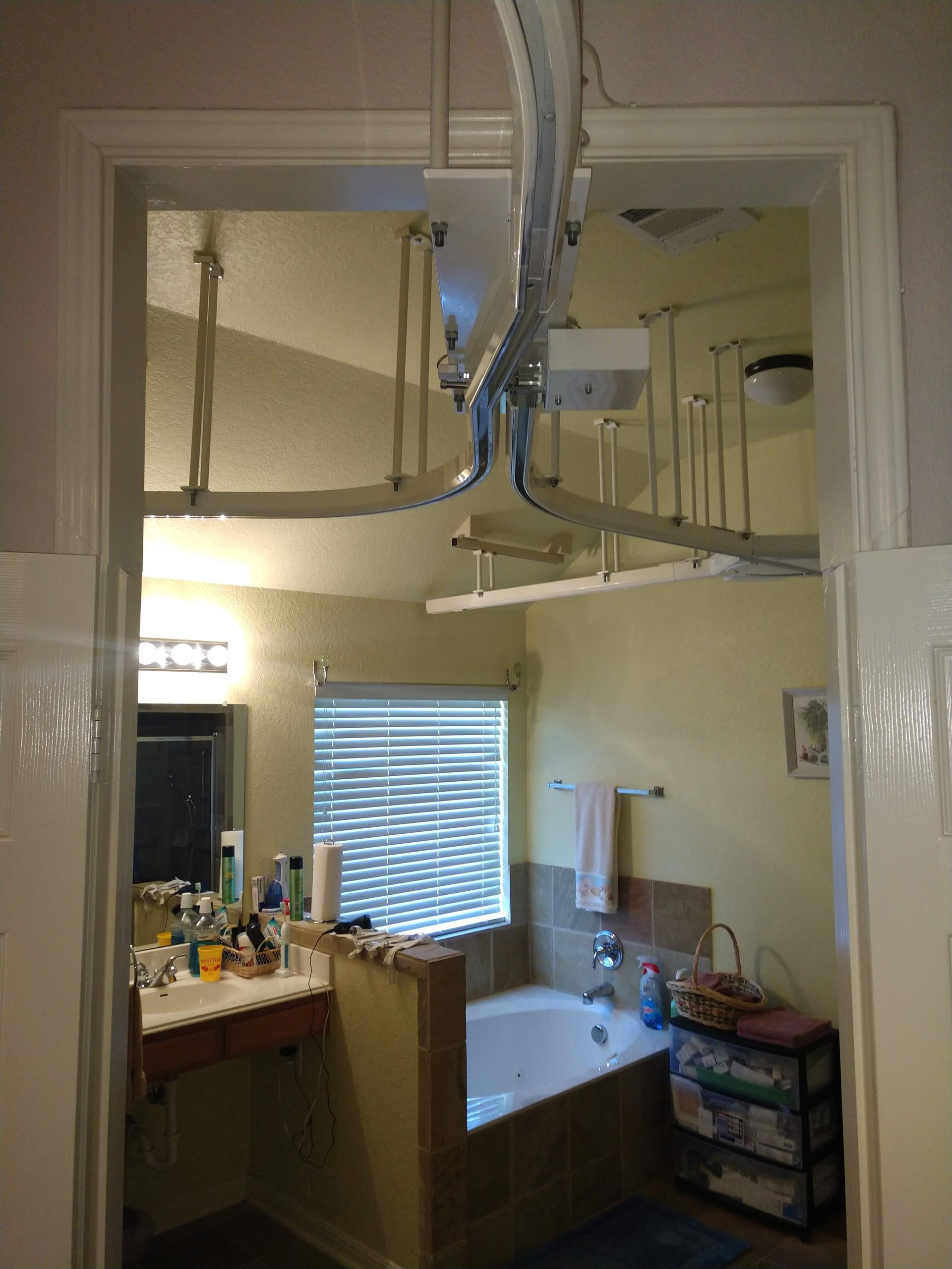 View from a doorway into a yellow-walled bathroom, showing a bathtub, sink, and overhead lift track.