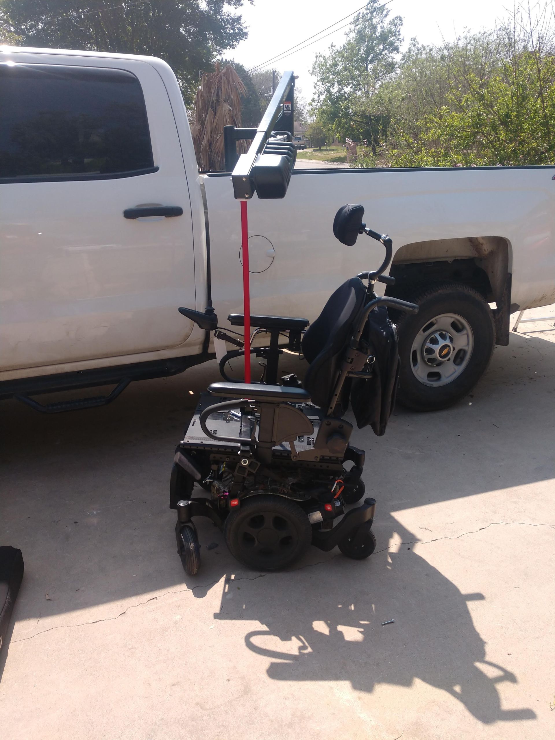 Black power wheelchair with a lift arm attached to the back, parked in front of a white truck.