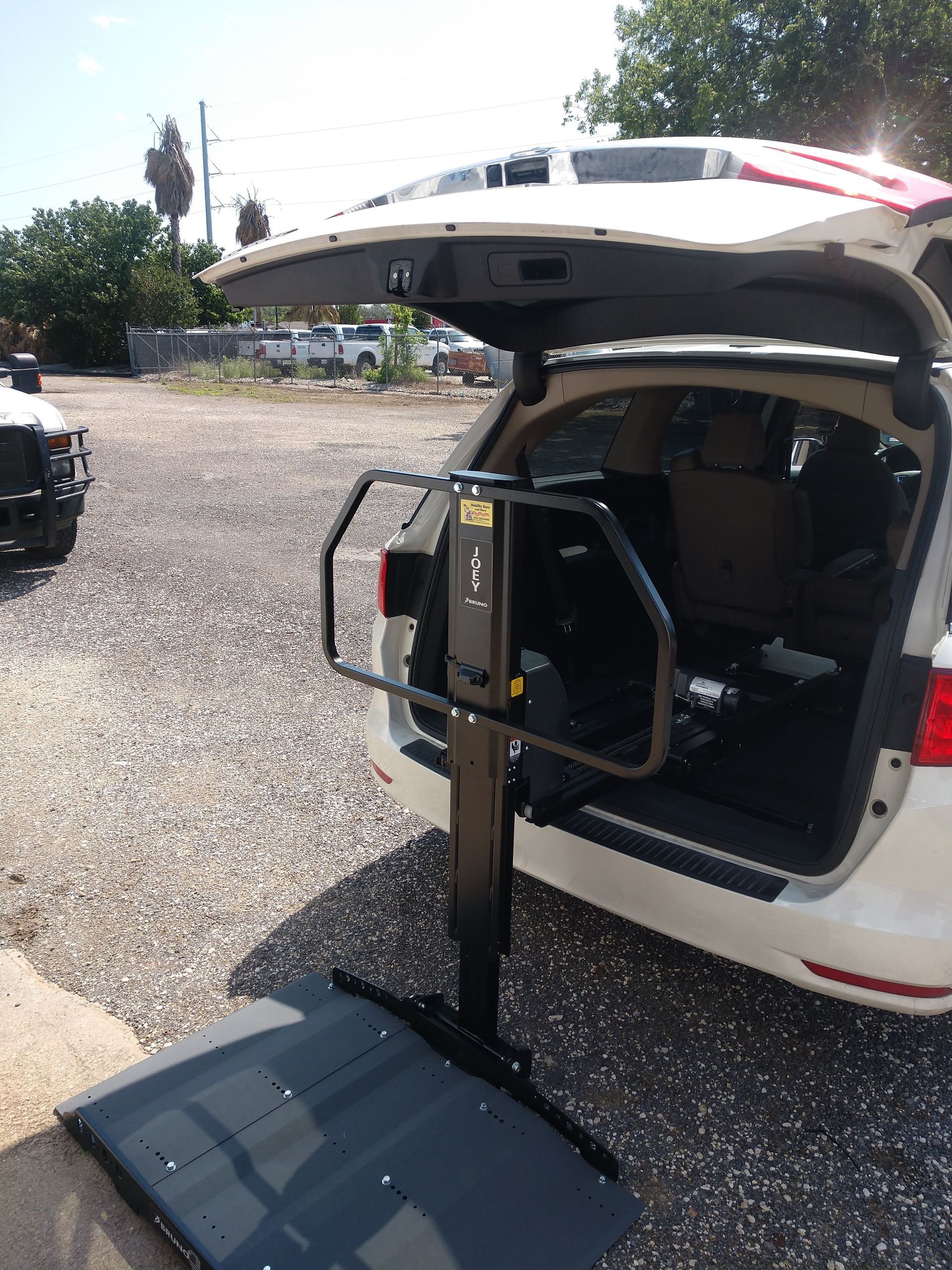 A white minivan with a wheelchair lift deployed, parked on a gravel lot, in front of an open hatch.