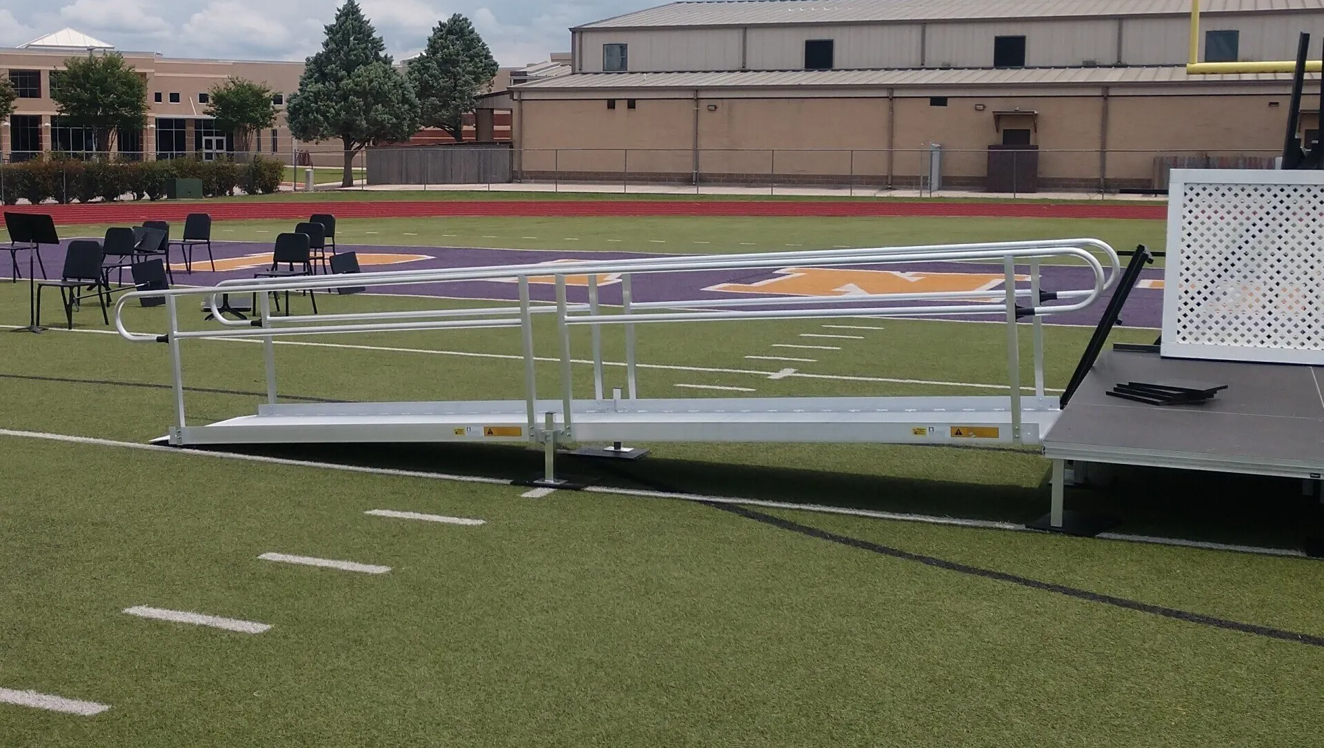 A metal ramp on a football field. Purple and white markings on the grass. Chairs and a building in the background.