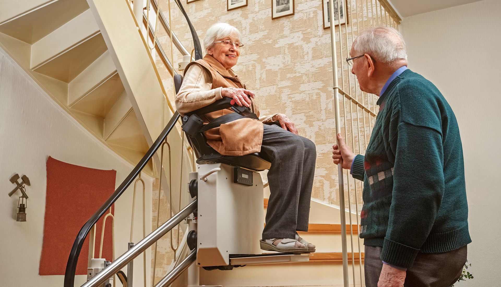An elderly woman smiles as she rides a stairlift down, a man standing beside her.