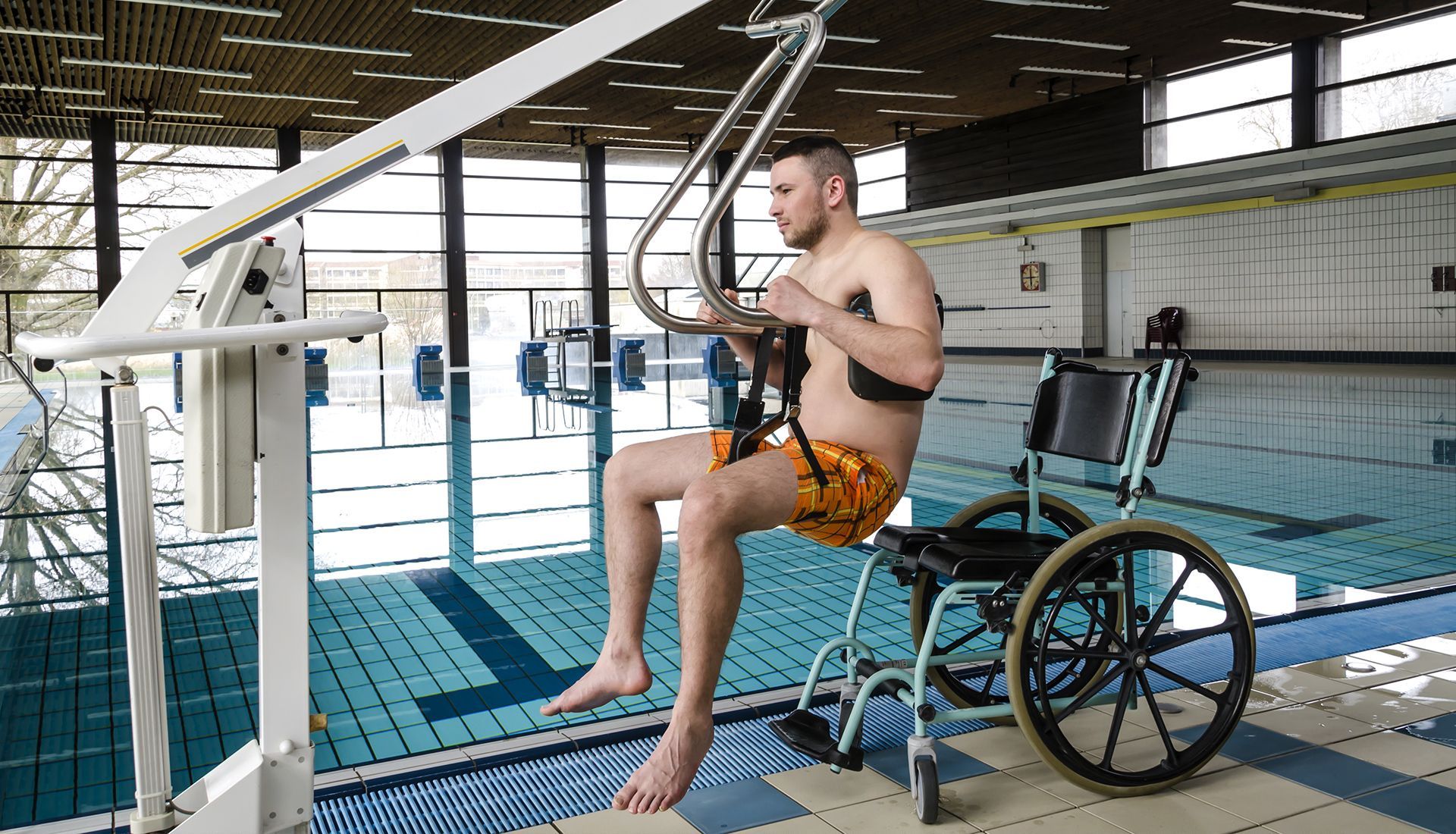 Man using a pool lift to transfer from wheelchair into a swimming pool. He is wearing swim trunks, and the pool is indoors.