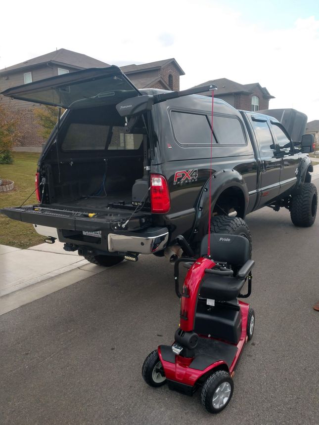 Black truck with open bed and canopy, red mobility scooter parked on the street.