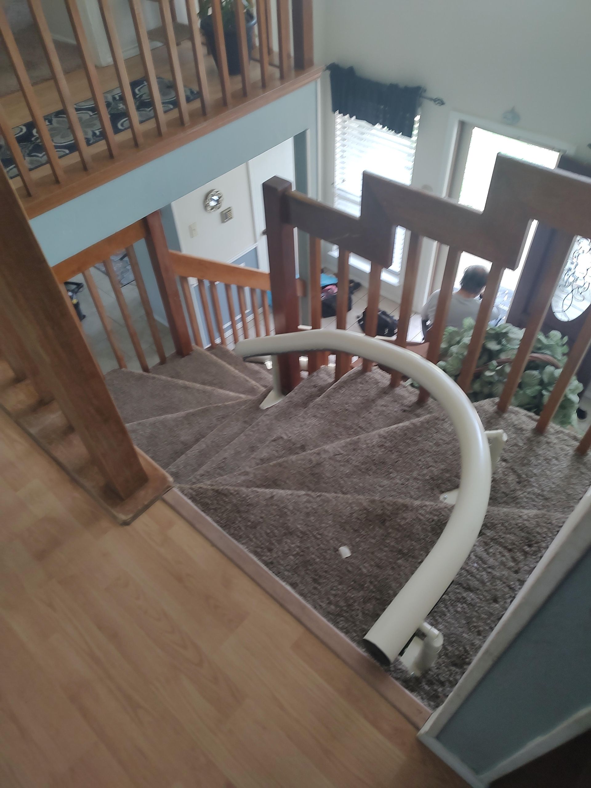 Staircase with a curved stairlift, wooden banisters, tan carpet. View from above, looking down.