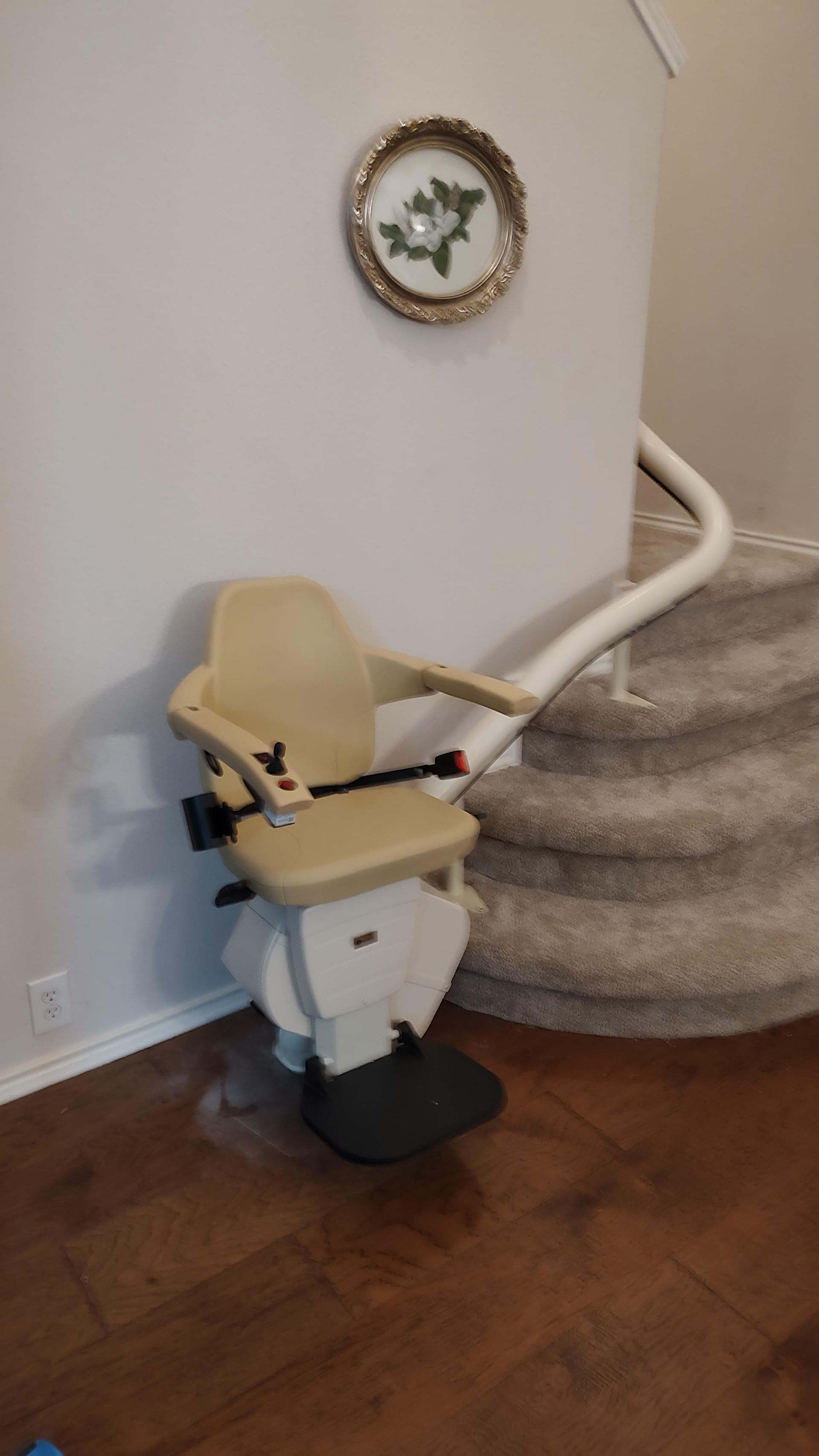 Beige stair lift next to a staircase, mounted on a white wall. A small, round picture hangs above.