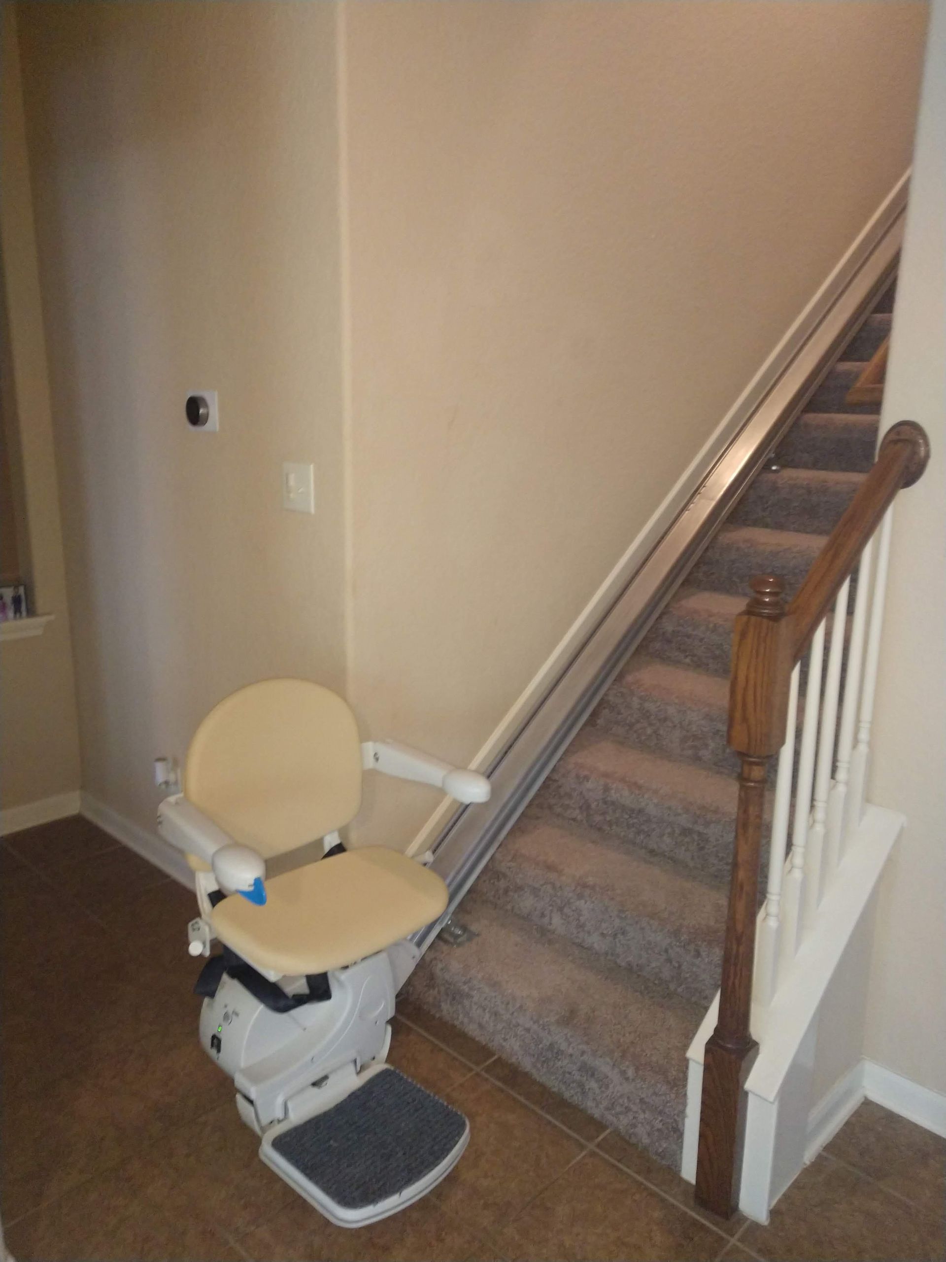 Stairlift on a beige wall, carpeted stairs, wooden railing, beige chair seat, brown stairs, and a white banister.