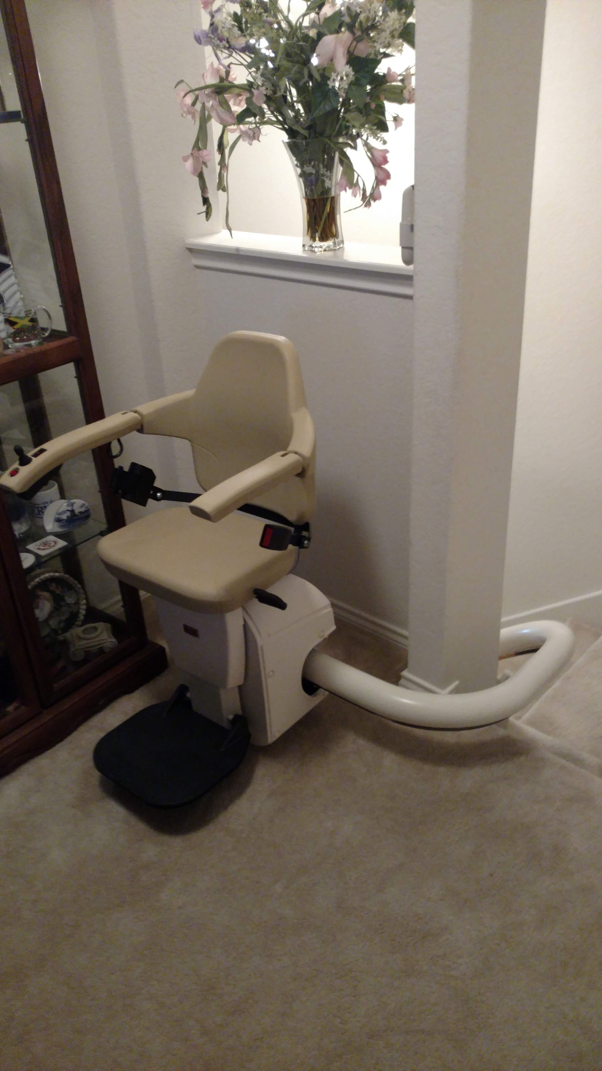 Cream-colored stairlift chair against a white wall. Carpeted floor, dark footrest.