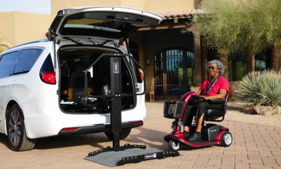 Woman on a red scooter using a vehicle lift to enter a white minivan. Desert home in background.