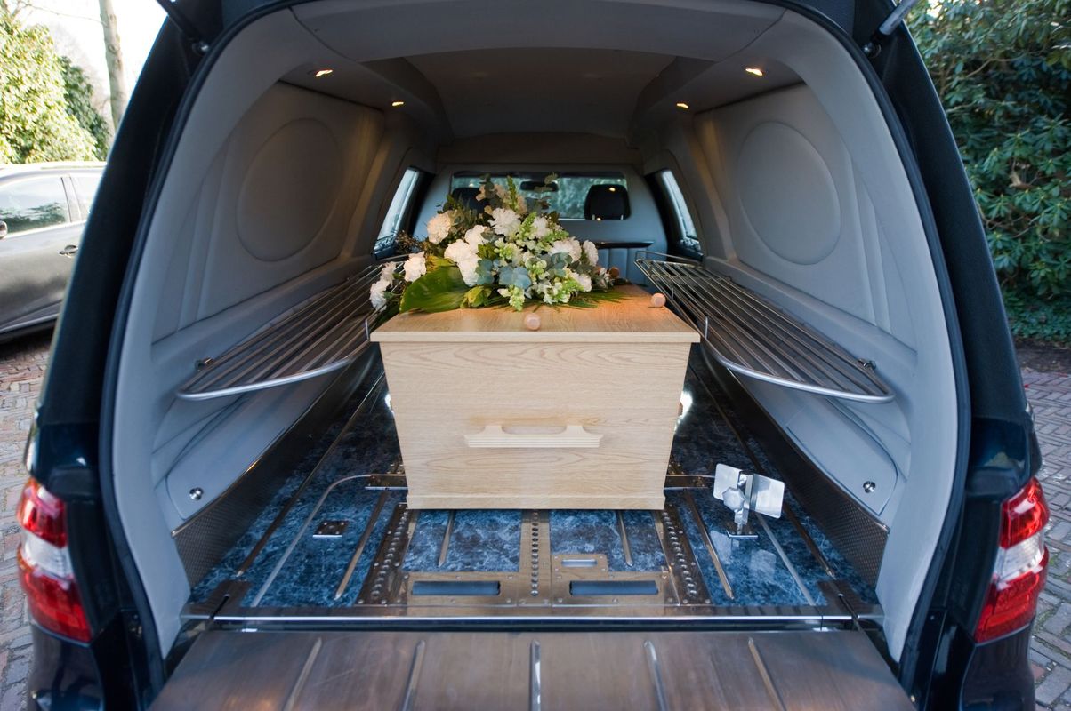 Una bara di legno per fiori all'interno del vano posteriore aperto di un carro funebre, con una composizione floreale sopra.