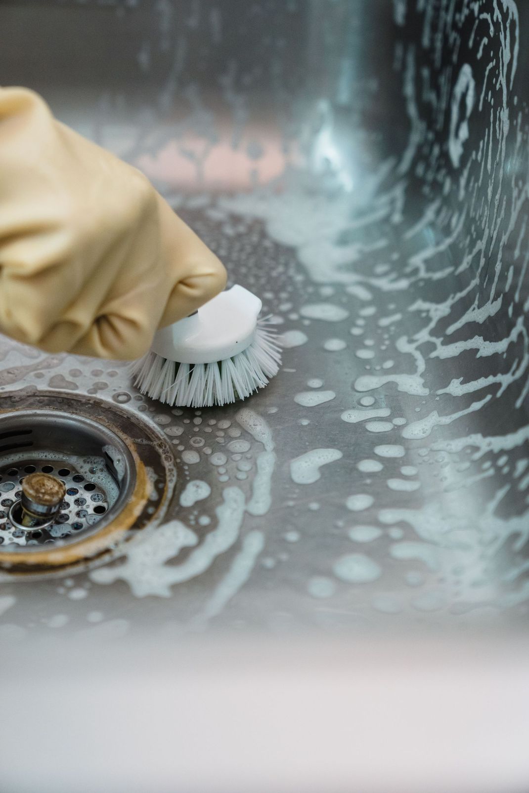 Gloved hand scrubbing a stainless steel sink drain with a brush and soap suds