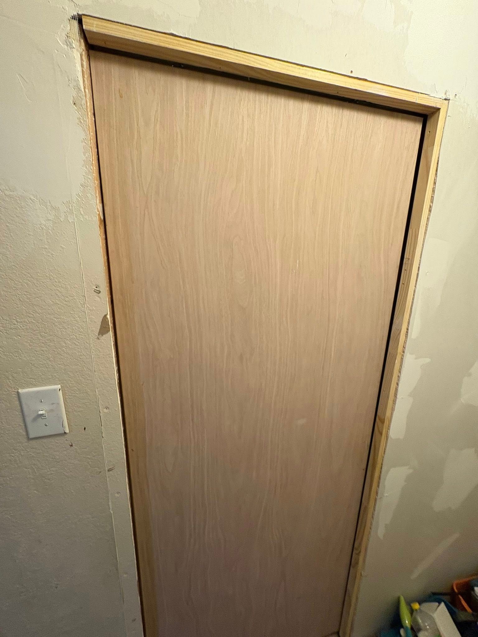A wooden door in a wall opening, trimmed with light wood. A light switch is visible on the left.