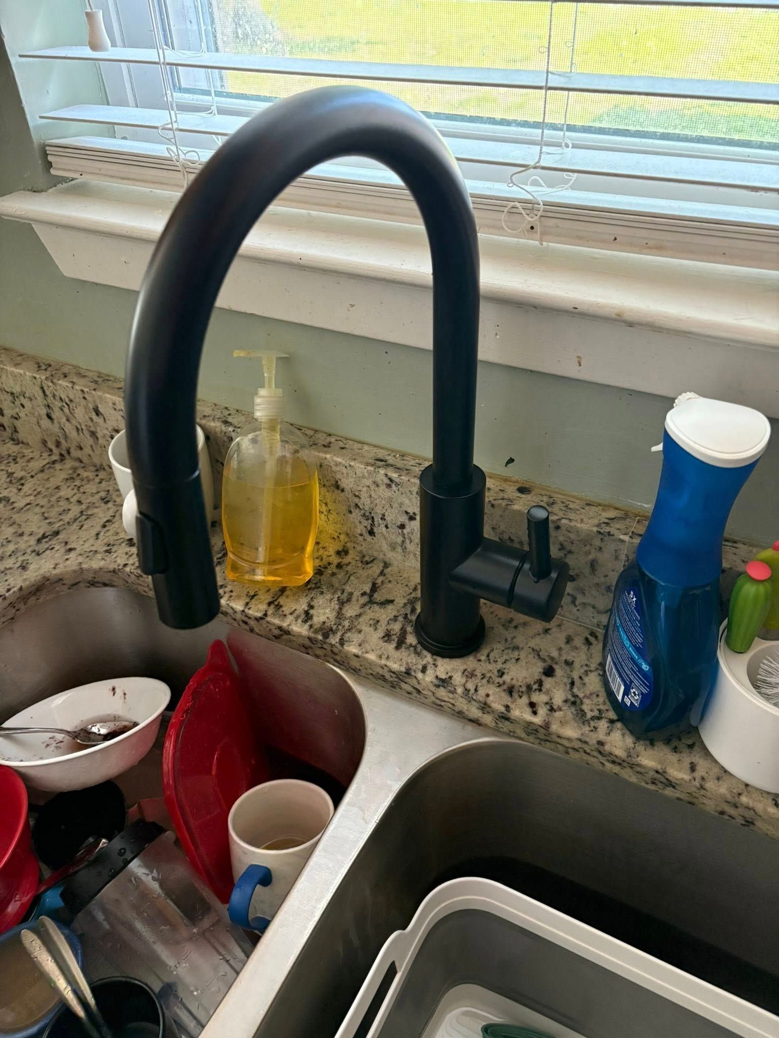 Black kitchen faucet over a dirty sink with dishes. A soap dispenser and blue bottle sit nearby.