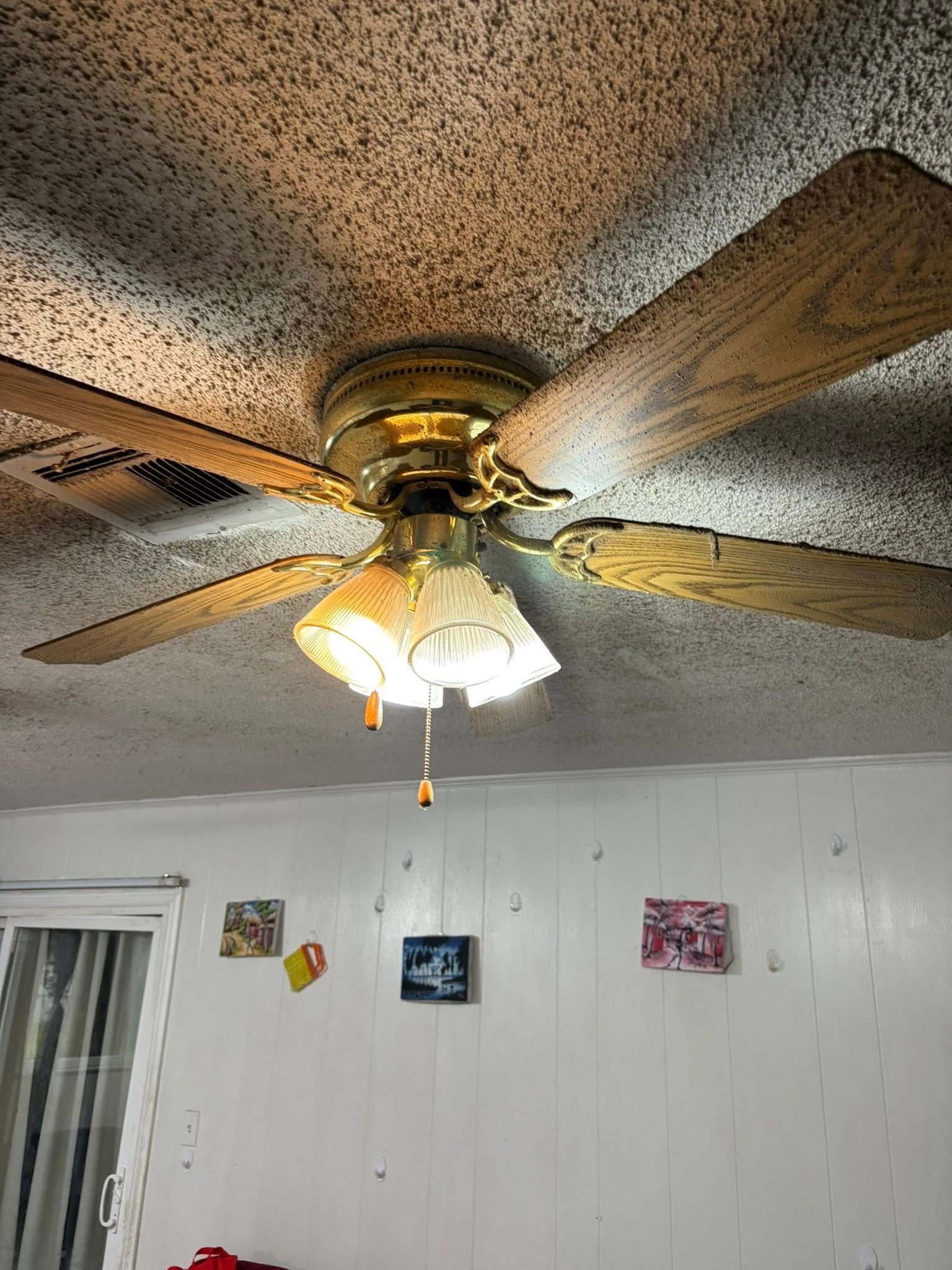 Ceiling fan with gold finish and wooden blades, a light fixture hangs below, mounted on a textured ceiling in a room.
