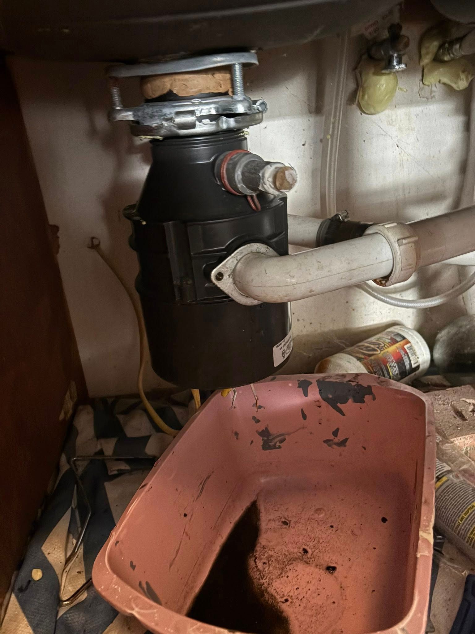 Garbage disposal under a sink with attached drainpipe and electrical cord. A pink plastic basin sits below.