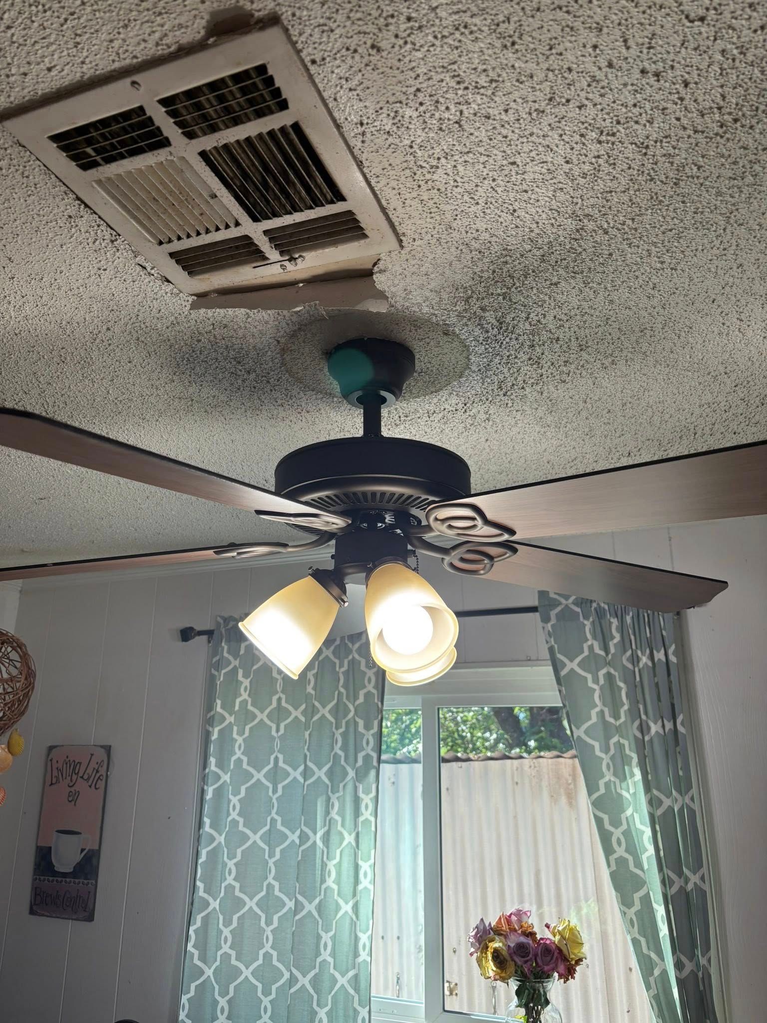 Ceiling fan with lights below a vent in a room with light blue curtains and a window.