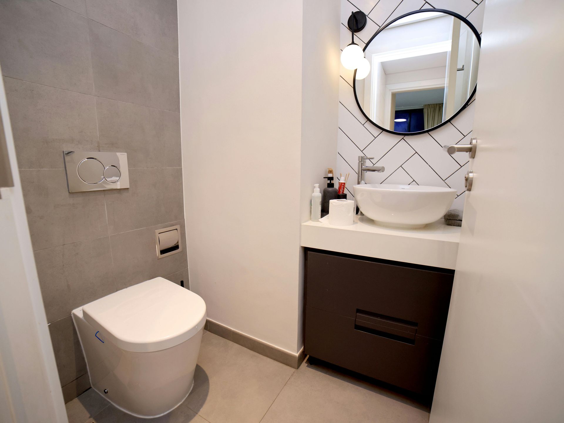 Small modern bathroom with white and gray tiles, toilet, sink, mirror, and dark brown cabinet.