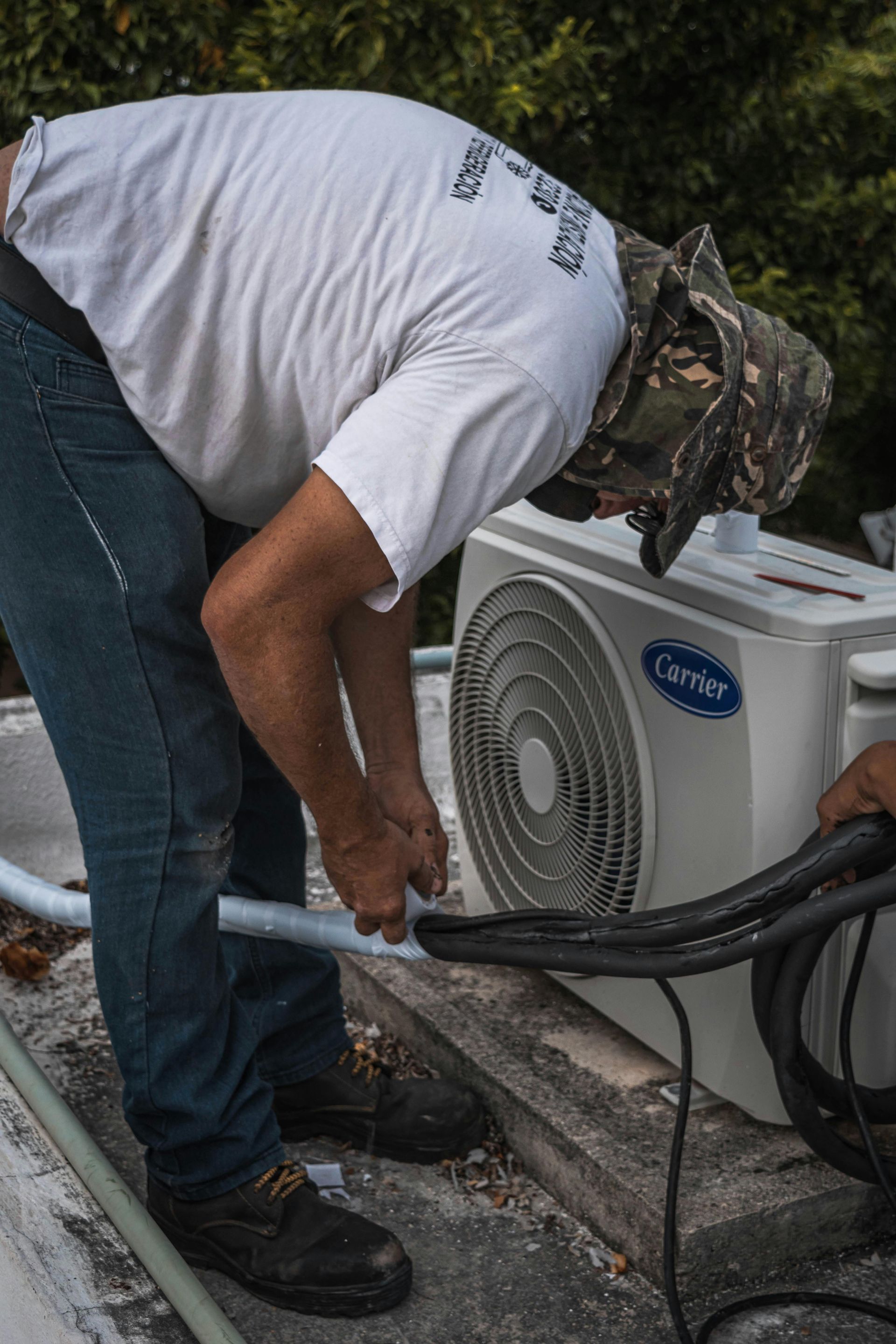 Man in white shirt and camo hat working on an outdoor Carrier air conditioning unit, on a rooftop.