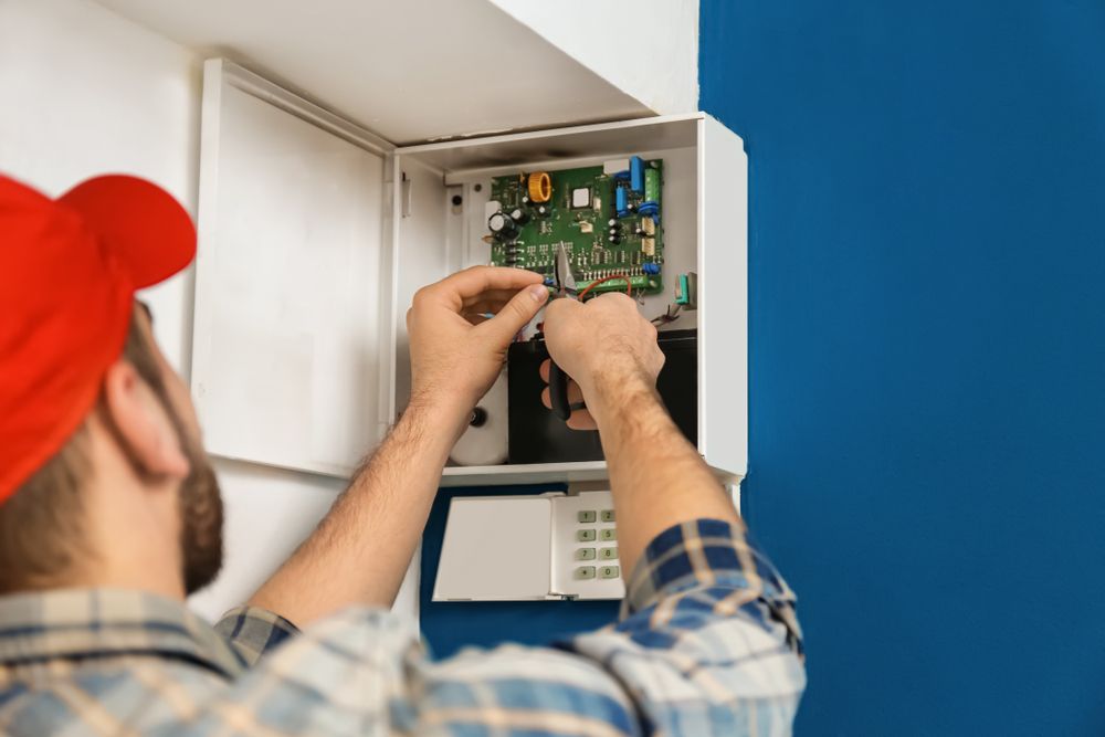 Technician in a Red Cap Repairs a Security System Panel Mounted on a Blue Wall — Holders Electrical In Tenterfield, NSW