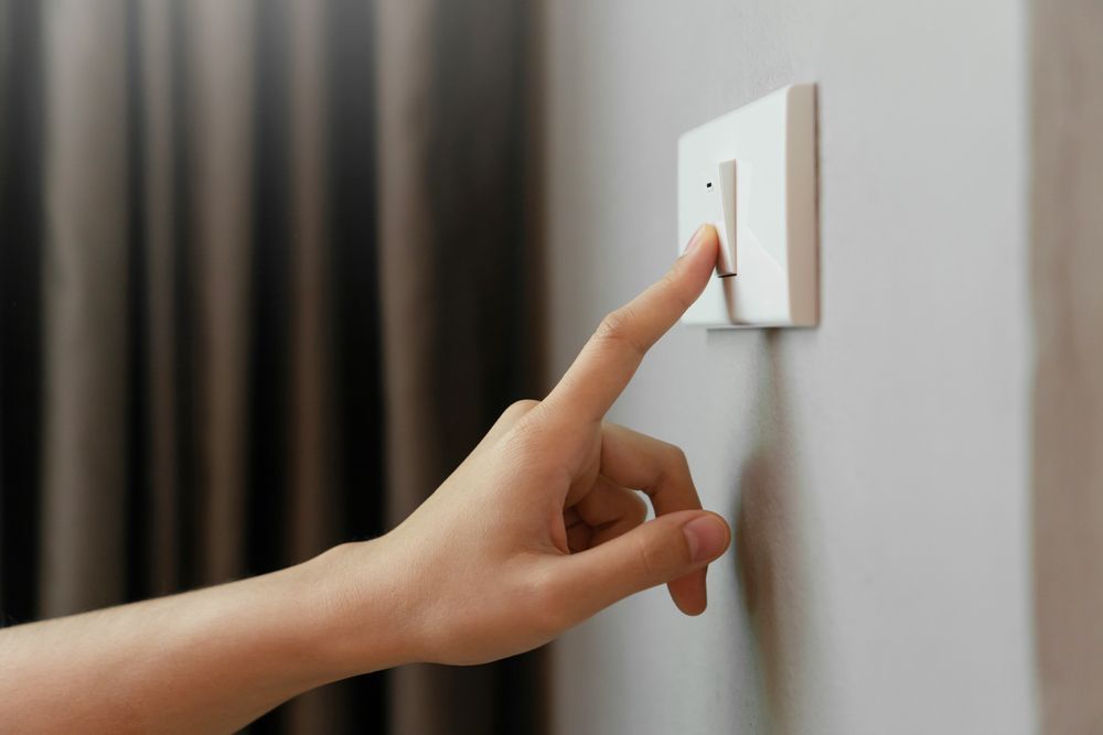 Hand pressing a light switch on a white wall. Grey curtain in the background — Holders Electrical In Warwick, QLD