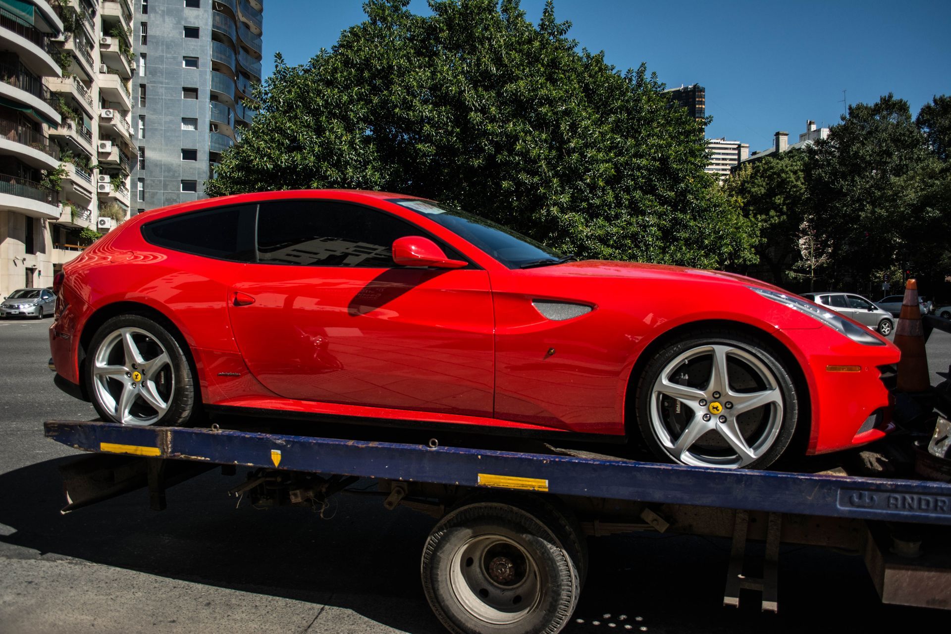 Red sports car on a flatbed tow truck outdoors beside apartment buildings and trees.