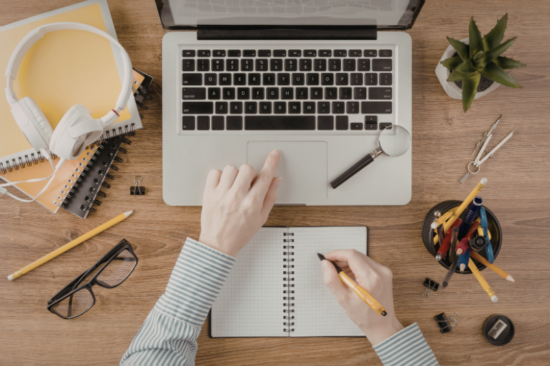 Overhead view of a workspace with a laptop, a hand writing in a notebook, headphones, a plant, and stationery.