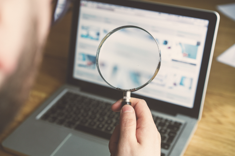 Hand holding a magnifying glass over a laptop screen, suggesting close inspection or search.