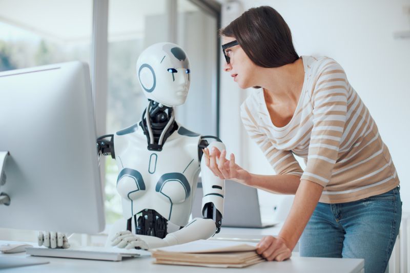 Woman in glasses gestures to robot at a computer, in a modern office setting.