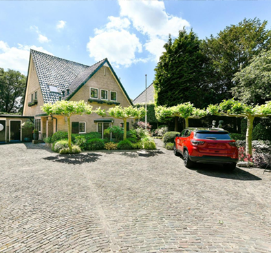 Red car parked in front of a house with a brick driveway and landscaped yard, under a sunny sky.