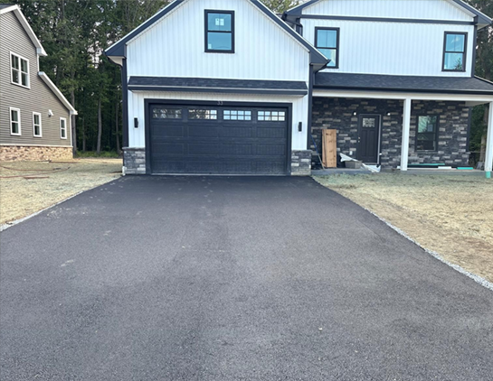 Black asphalt driveway leading to a two-story house with a black garage door and white siding.