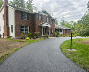 Newly paved asphalt driveway leading to a two-story brick house with green lawn and trees.