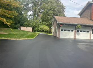 Newly paved black driveway leads to a brick building with white garage doors, set amid green lawn and trees.
