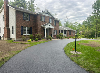 Two-story brick house with newly paved driveway. Green lawn, trees in background, cloudy sky.