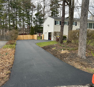 Black asphalt driveway leading to a two-story house with a garage, trees and a wooden fence.