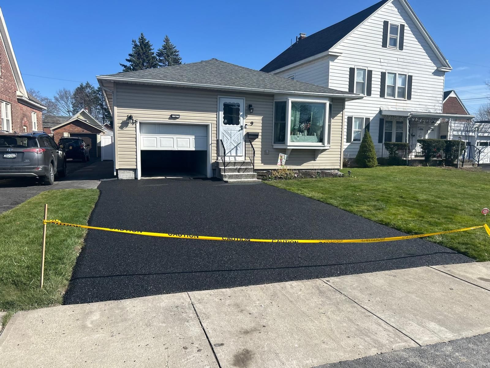 Freshly paved black driveway in front of a tan house with a garage and bay window, yellow caution tape across the lawn.