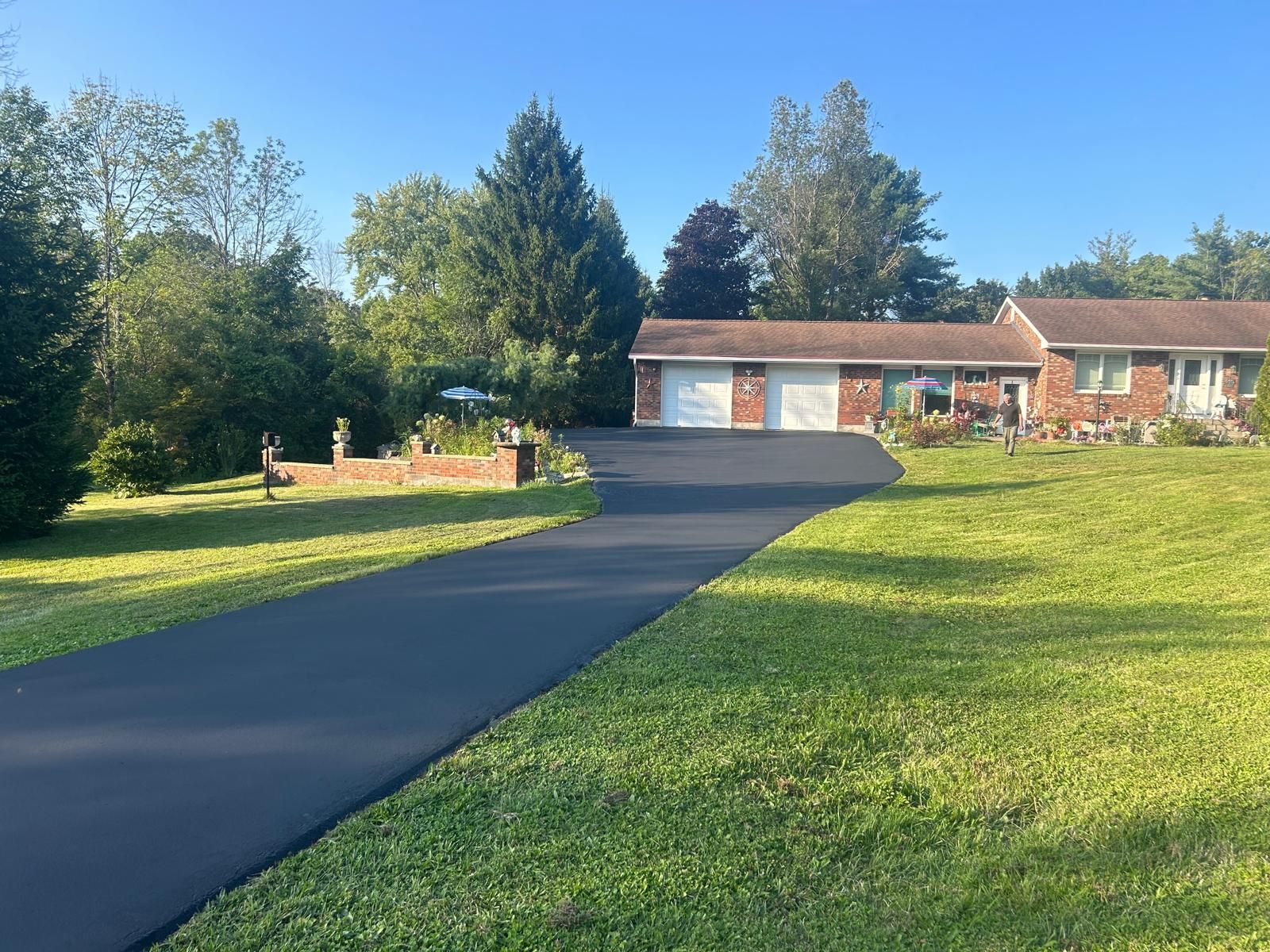 Black paved driveway leading to a brick house with a two-car garage, on a green lawn under a blue sky.