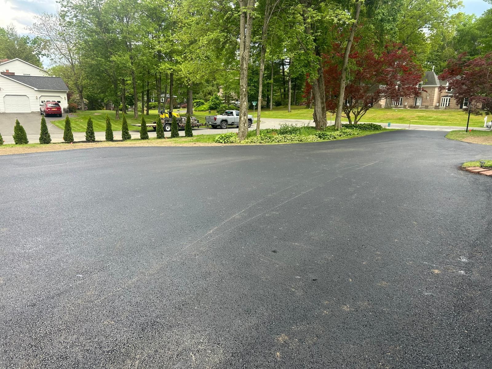Black asphalt driveway in front of a house, lined with small trees and other landscaping.