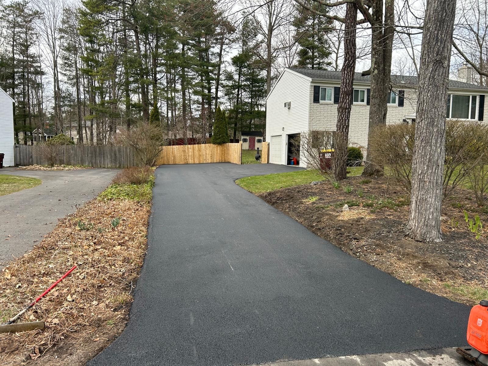 Newly paved asphalt driveway leading to a two-story house, bordered by trees and a wooden fence.