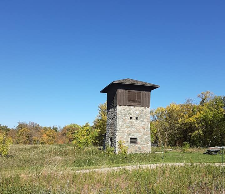 A stone-based observation tower with a brown wooden top stands in a grassy field under a clear blue sky.