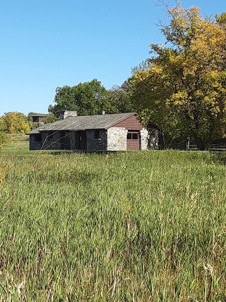 A historic stone house sits in a tall grassy field under a clear blue sky, surrounded by trees with autumn foliage.