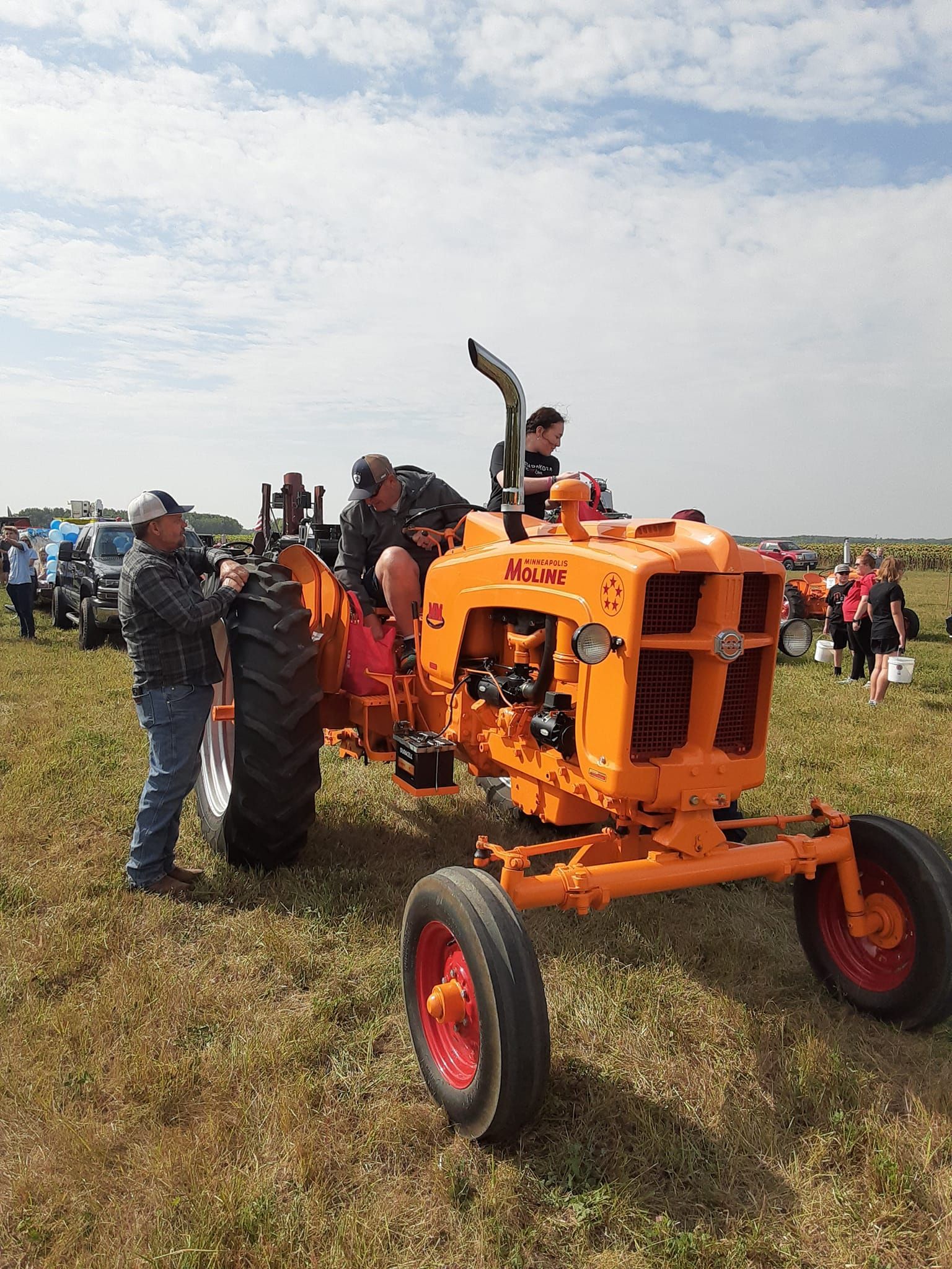 Orange Allis-Chalmers tractor with people on it, in a field, at an outdoor event.