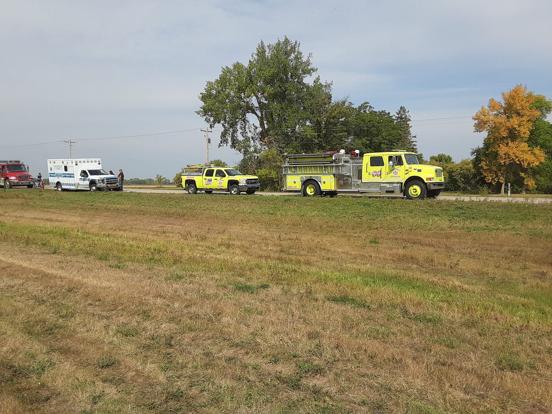 Emergency vehicles on a roadside: fire truck, ambulance, and other response vehicles in a field under a cloudy sky.