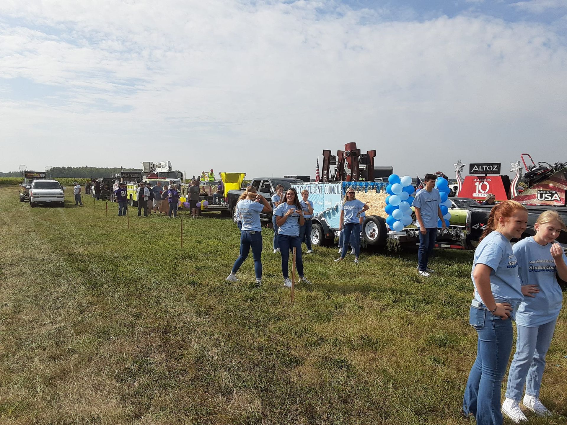 People gather near decorated floats in a field, under a cloudy sky.